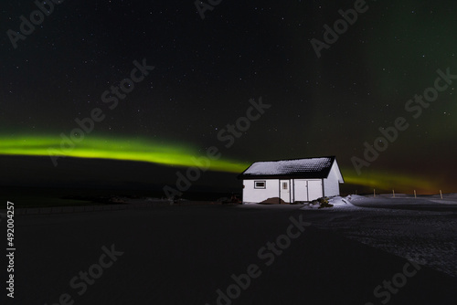 Mały skandynawski dom w nocy oświetlony piękną zorzą polarną, zielona zorza polarna przecina ciemne nocne niebo nad Lofotami w Norwegii