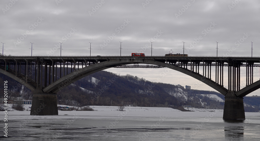 Landscape with a span of a large arched bridge and a frozen river. A tram and a bus have met on the bridge and cannot separate. A tram with an empty passenger compartment is moving over a beautiful br