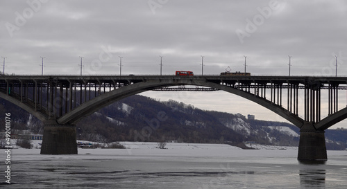 Fototapeta Naklejka Na Ścianę i Meble -  Landscape with a span of a large arched bridge and a frozen river. A tram and a bus have met on the bridge and cannot separate. A tram with an empty passenger compartment is moving over a beautiful br