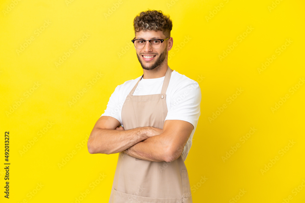 Restaurant waiter blonde man isolated on yellow background with arms crossed and looking forward Restaurant waiter blonde man isolated on yellow background with arms crossed and looking forward