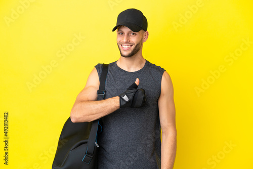 Young sport blonde man with sport bag isolated on yellow background giving a thumbs up gesture