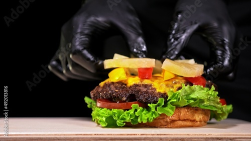 Craft burger is cooking on black background in black food gloves. Consist: red sauce salsa, lettuce, red onion, pickle, cheese, chilli green pepper, air bun and marbled meat beef. Not made ideal
