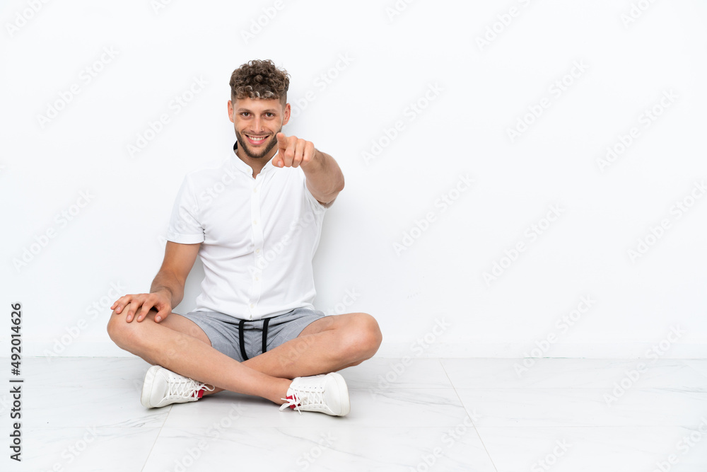 Young blonde man sitting on the floor isolated on white background points finger at you with a confident expression Young blonde man sitting on the floor isolated on white background points finger at you with a confident expression