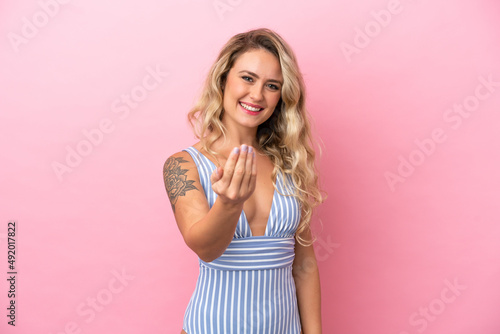 Young Brazilian woman in swimsuit in summer holidays isolated on pink background inviting to come with hand. Happy that you came