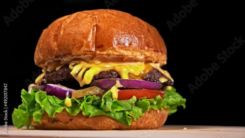 Craft burger is cooking on black background in black food gloves. Consist: sauce, lettuce, tomato, red onion, pickle, cheese, bacon, air bun and marbled meat beef. Not made ideal. Looks real, loving