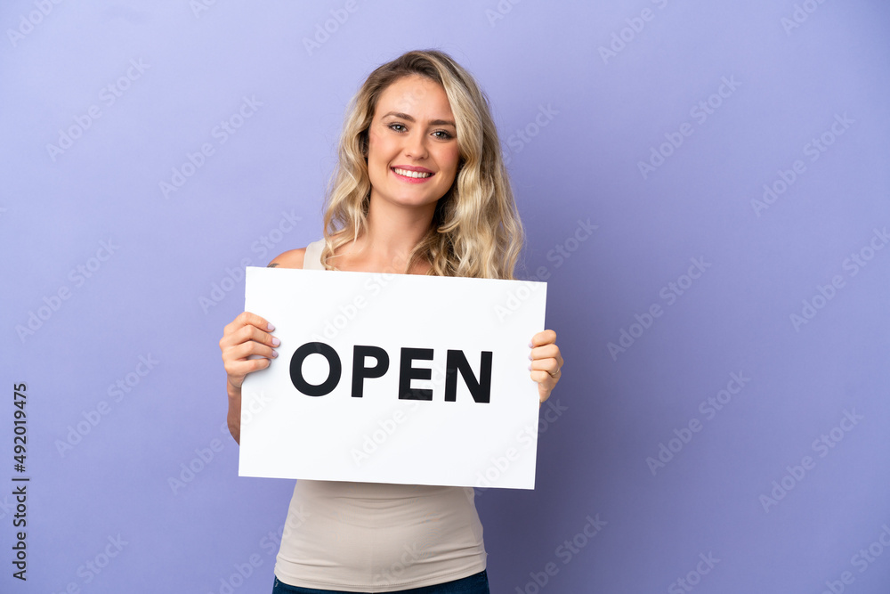 Young Brazilian woman isolated on purple background holding a placard with text OPEN with happy expression Young Brazilian woman isolated on purple background holding a placard with text OPEN with happy expression