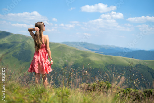 Young happy woman traveler in red dress standing on green grassy hillside on a windy day in summer mountains enjoying view of nature