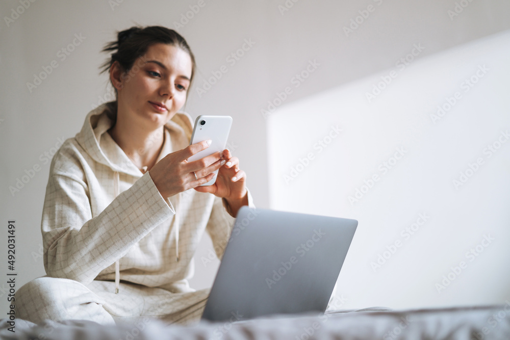Beautiful smiling woman teenager girl student freelancer with dark long hair in casual using mobile phone working on laptop sitting on couch at the home