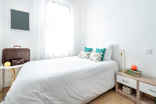 King size bed with white and green cushions, lamp without lampshade on bedside table and vintage brown travel suitcase