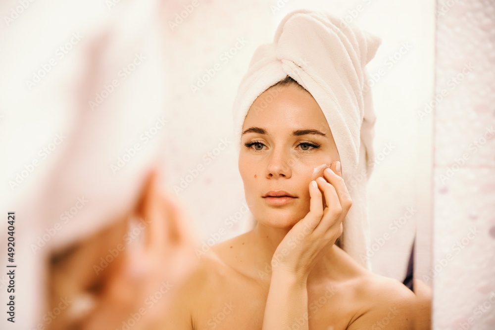 Young beautiful woman using face cream moisturizing lotion after bath. Pretty attractive girl in a towel on her head stands in front of a mirror in a home bathroom. Daily hygiene and skin care Young beautiful woman using face cream moisturizing lotion after bath. Pretty attractive girl in a towel on her head stands in front of a mirror in a home bathroom. Daily hygiene and skin care