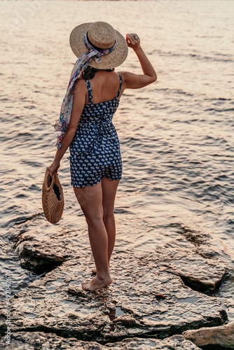 A woman in a dress, hat and with a straw bag is standing on the beach enjoying the sea. Happy summer holidays