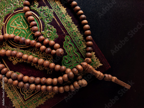 Holy Al Quran with written arabic calligraphy meaning of Al Quran and rosary beads or tasbih on black background.