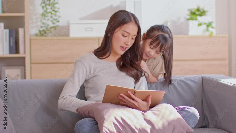 An Asian mom and her kid are reading together. Mom is teaching her child.child is concentrate on her mom.Mother is sitting on the sofa and her daughter is standing behind the sofa.Mom and kid concept An Asian mom and her kid are reading together. Mom is teaching her child.child is concentrate on her mom.Mother is sitting on the sofa and her daughter is standing behind the sofa.Mom and kid concept