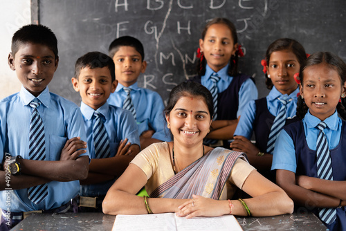 Happy Teacher with students around looking camera at classroom in front of black board - concept of support, education, growth, employment and job.