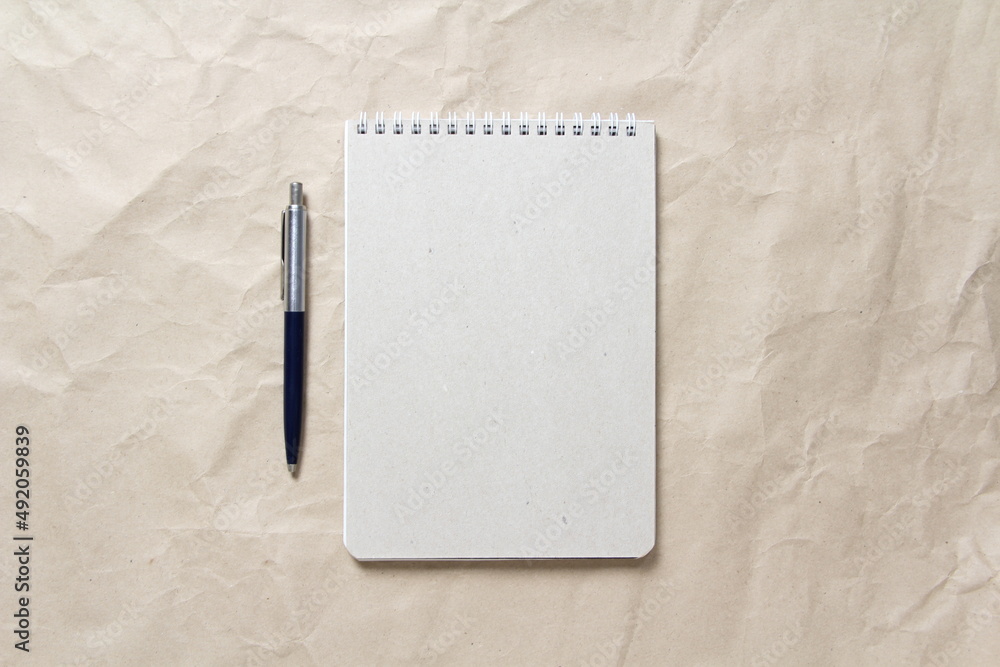 Gray notepad with white coiled spring and pen on a background of beige crumpled craft paper. With empty space for text and design Gray notepad with white coiled spring and pen on a background of beige crumpled craft paper. With empty space for text and design