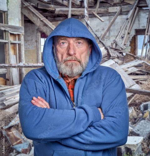 portrait of sad and gloomy senior man in a hoodie against rubbish of a ruined house