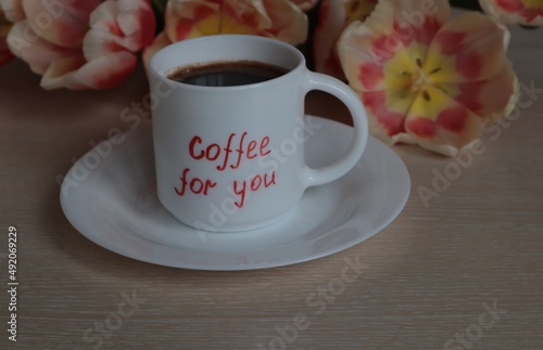 A white mug with black coffee stands against a background of tulips on a light surface.