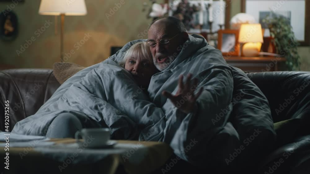 Upset elderly man discussing gas crisis and war with sad wife while wrapping in warm blanket on couch in dim cold living room at home Upset elderly man discussing gas crisis and war with sad wife while wrapping in warm blanket on couch in dim cold living room at home