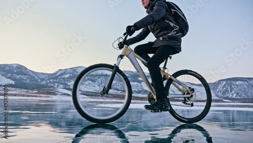 Fototapeta Naklejka Na Ścianę i Meble -  Man is riding bicycle on the ice. Ice of the frozen Lake Baikal. Teenage is dressed in black down jacket, cycling backpack, helmet. Tires on covered with special spikes. Traveler boy is ride cycle.