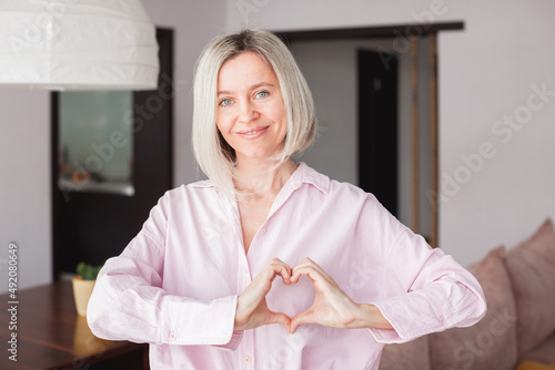 Middle-aged woman sit in living room connected fingers showing heart symbol