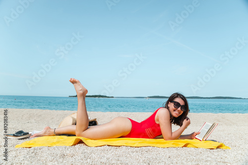 young sexy stunning woman in red swimsuit reading book on sea beach