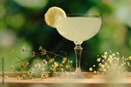 Drinking glass of daiquiri cocktail and summer flowers on wooden desk outdoors. Summer refreshing alcoholic citrus cocktail. photo