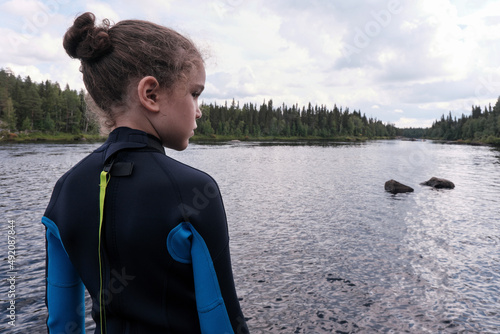 Portrait of cute curly boy in a wetsuit on the river bank on a cloudy summer day. photo