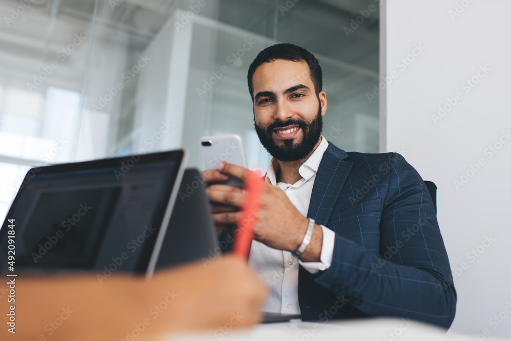 Portrait of cheerful male boss with modern cellular device smiling at camera during working day in office interior, happy Middle Eastern corporate director with smartphone enjoying business lifestyle Portrait of cheerful male boss with modern cellular device smiling at camera during working day in office interior, happy Middle Eastern corporate director with smartphone enjoying business lifestyle