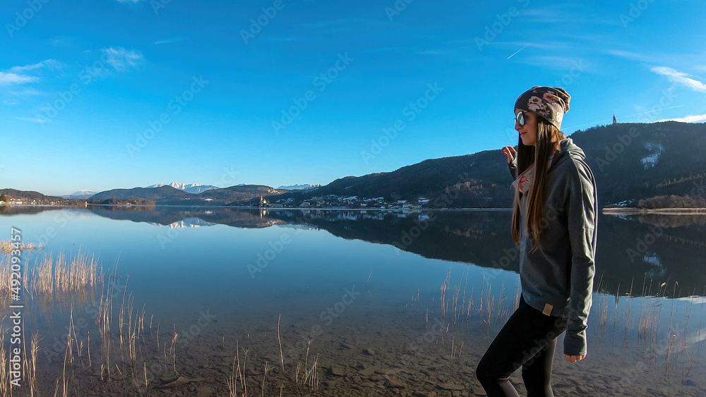 Naklejka premium Happy woman walking along the shore of beautiful Woerthersee in Poertschach, Carinthia, Austria. Scenic lake landscape surrounded by Alps. Serenity. Fresh and clean air. Reflection in Lake Woerth