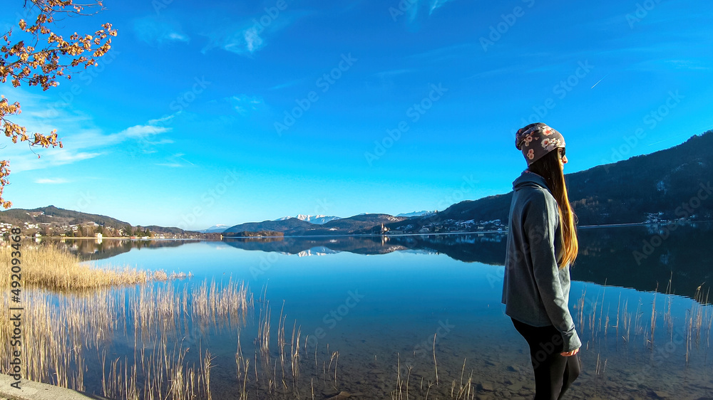 Fototapeta premium Happy woman walking along the shore of beautiful Woerthersee in Poertschach, Carinthia, Austria. Scenic lake landscape surrounded by Alps. Serenity. Fresh and clean air. Reflection in Lake Woerth