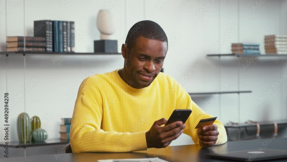 Concentrated financial director in yellow shirt looks at bank card in hand and enters digits to smartphone Concentrated financial director in yellow shirt looks at bank card in hand and enters digits to smartphone