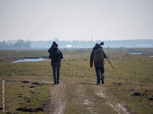 Fototapeta Birdwatchers with big cameras and backpacks walking in polder meadow landscape