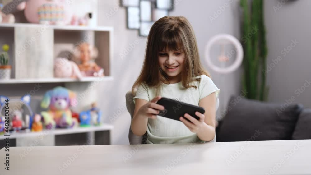 Girl Playing in Video Game on His Smartphone at the Desk at Home in the Living Room. Cute Small Kid Girl Using Digital Computer Phone Playing Online Games Girl Playing in Video Game on His Smartphone at the Desk at Home in the Living Room. Cute Small Kid Girl Using Digital Computer Phone Playing Online Games