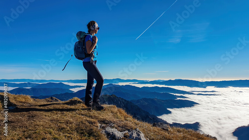 A woman enjoying the panoramic view from mount Eisenerzer Reichenstein in Styria, Austria, Europe. The Ennstal valley is covered in clouds and fog. Hiking trail, Wanderlust. Sunny day. Freedom concept