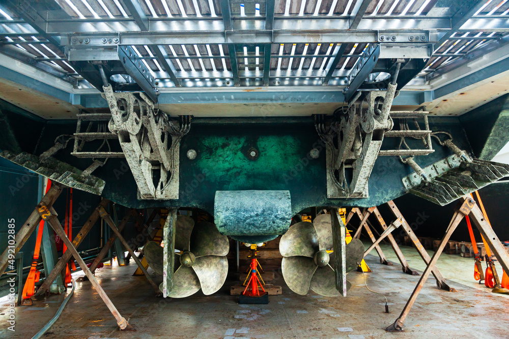 The back of a large motor yacht, standing on wooden blocks in a dry dock. Two propellers and zinc protective inserts.