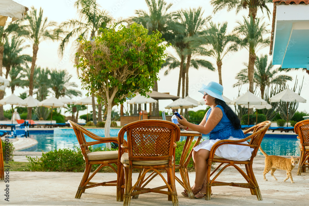 A young woman in a hat at a pool table among palm trees drinks a cocktail and works remotely on a laptop.