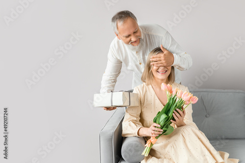 An attractive old man gives his charming wife flowers and a gift for the holiday on March 8. Surprise with eyes closed.