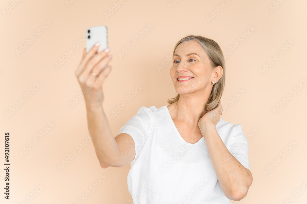 Old attractive woman takes a selfie on a beige background. Isolated portrait in light clothes. Old attractive woman takes a selfie on a beige background. Isolated portrait in light clothes.