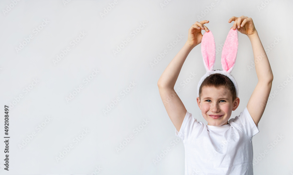 Banner of funny playful boy is wearing bunny ears and white t shirt on background of white wall during Easter holiday. Preparing for the holiday is a joy for kids. Little child celebrate Easter Banner of funny playful boy is wearing bunny ears and white t shirt on background of white wall during Easter holiday. Preparing for the holiday is a joy for kids. Little child celebrate Easter