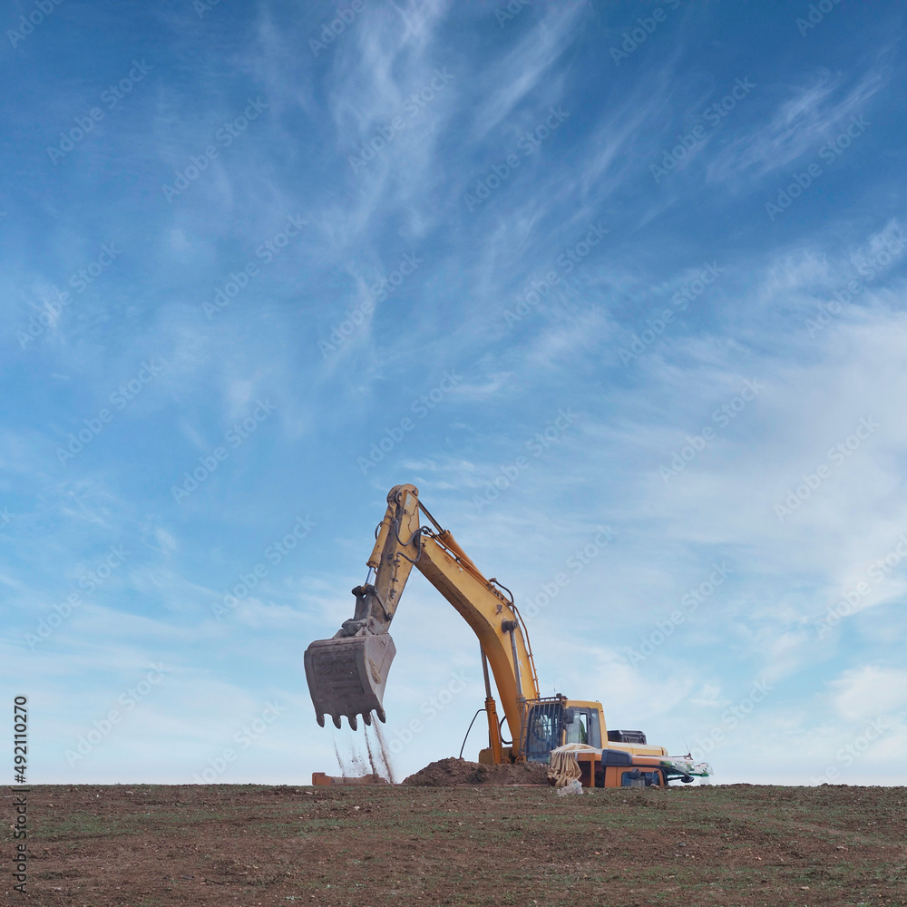Naklejka premium excavator working moving earth and loading on a truck with blue sky and square format clouds