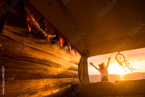 Overjoyed tourist jumping against a golden sunset light viewed from inside a wooden hand made classic van camper. People and travel vanlife lifestyle. Freedom and happiness in summer holiday vacation photo