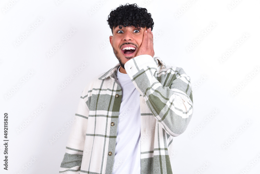 Shocked, astonished young arab man with curly hair wearing overshirt looking surprised in full disbelief wide open mouth with hand near face. Positive emotion facial expression body language. Shocked, astonished young arab man with curly hair wearing overshirt looking surprised in full disbelief wide open mouth with hand near face. Positive emotion facial expression body language.