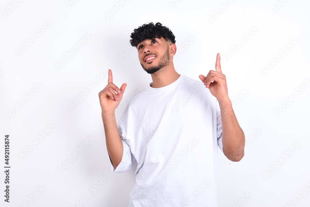 Successful friendly looking young arab man with curly hair wearing white t-shirt over white background exclaiming excitedly, pointing both index fingers up, indicating something. Successful friendly looking young arab man with curly hair wearing white t-shirt over white background exclaiming excitedly, pointing both index fingers up, indicating something.