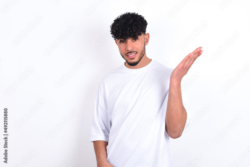 Studio shot of frustrated young arab man with curly hair wearing white t-shirt over white background gesturing with raised palm, frowning, being displeased and confused with dumb question. Studio shot of frustrated young arab man with curly hair wearing white t-shirt over white background gesturing with raised palm, frowning, being displeased and confused with dumb question.