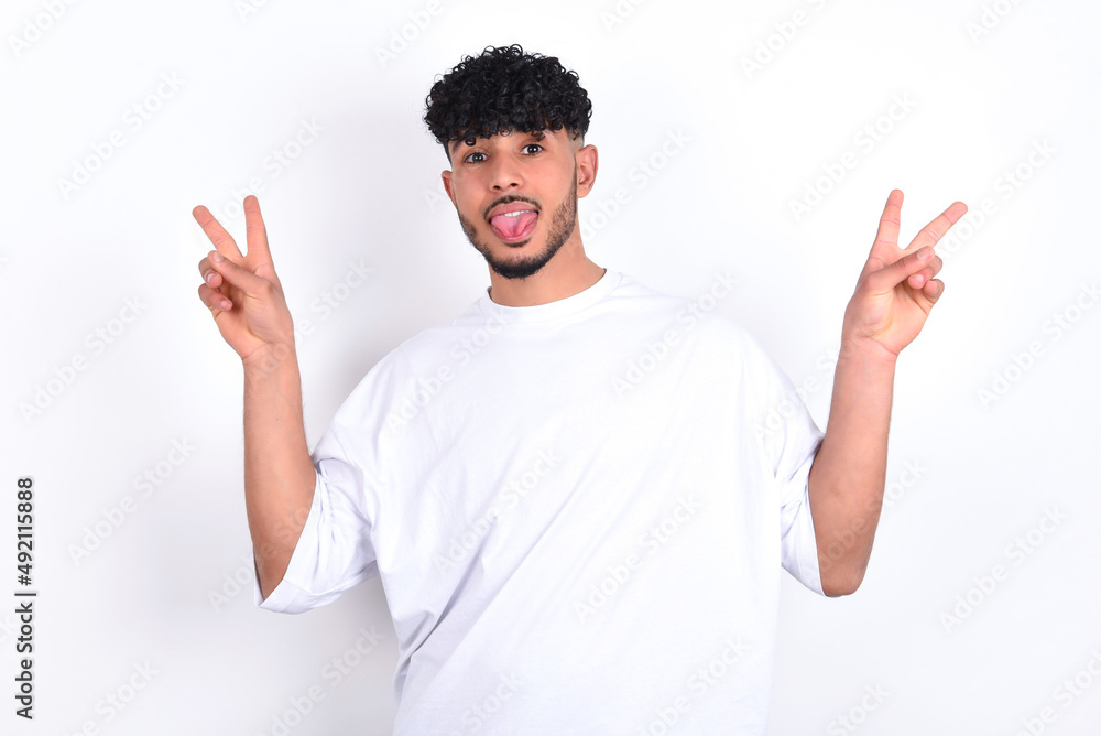 young arab man with curly hair wearing white t-shirt over white background with optimistic smile, showing peace or victory gesture with both hands, looking friendly. V sign. young arab man with curly hair wearing white t-shirt over white background with optimistic smile, showing peace or victory gesture with both hands, looking friendly. V sign.