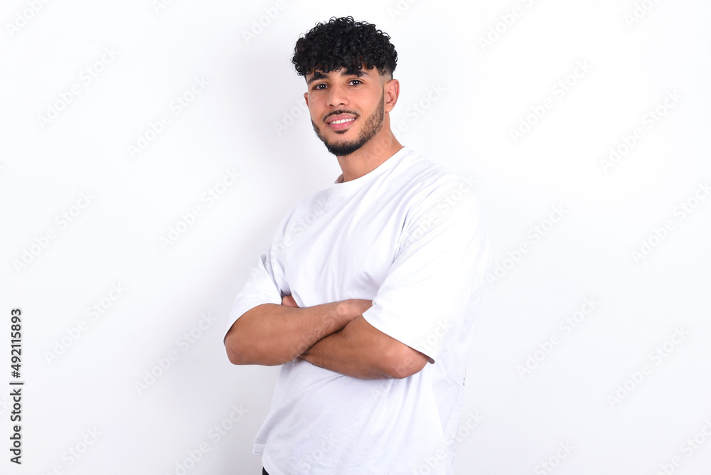 Image of cheerful young arab man with curly hair wearing white t-shirt over white background with arms crossed. Looking and smiling at the camera. Confidence concept. Image of cheerful young arab man with curly hair wearing white t-shirt over white background with arms crossed. Looking and smiling at the camera. Confidence concept.