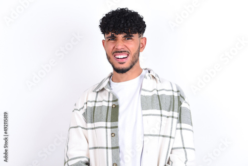 Mad crazy young arab man with curly hair wearing overshirt over white background clenches teeth angrily, being annoyed with coming noise. Negative feeling concept.