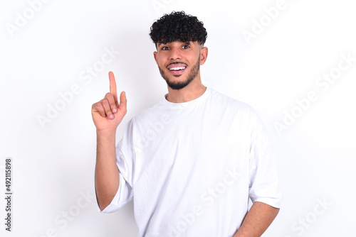 Pleasant looking young arab man with curly hair wearing white t-shirt over white background  has clever expression, raises one finger, remembers himself not to forget tell important thing. © Jihan