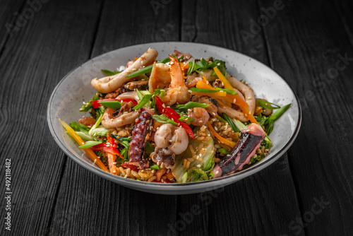 A plate of delicious rice with seafood: shrimp, squid, mussels and vegetables: carrots, sweet peppers, greens, spices and sauce on a black background