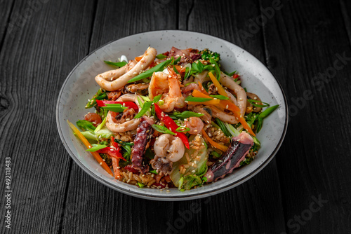 A plate of delicious rice with seafood: shrimp, squid, mussels and vegetables: carrots, sweet peppers, greens, spices and sauce on a black background